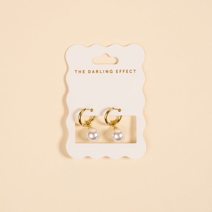 The Darling Effect’s Dainty Pearl Standout Earrings feature golden hoops with pearl drops, elegantly displayed on a scalloped white card—perfect for making a statement.