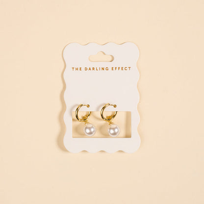 The Darling Effect’s Dainty Pearl Standout Earrings feature golden hoops with pearl drops, elegantly displayed on a scalloped white card—perfect for making a statement.