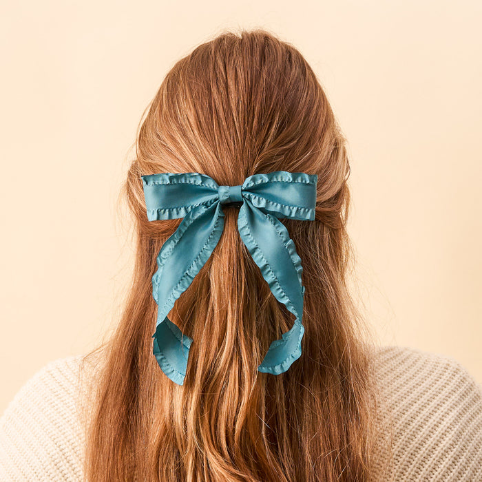 A person with long, wavy light brown hair, seen from behind, wears The Darling Effect's Dark Teal Ruffle Hair Bow and a cream knit sweater against a plain beige background.