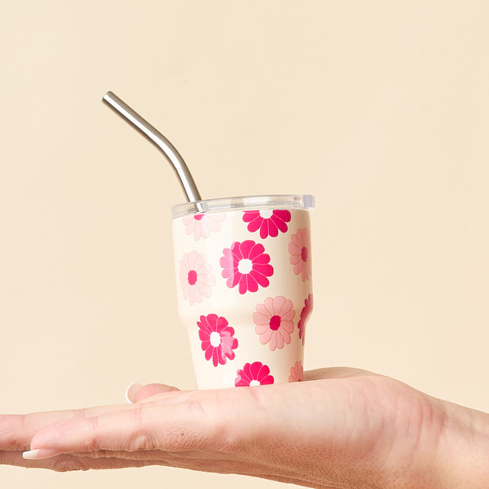 A hand holds the-darling-effect's Darling Daisy Hot Pink 2 oz Tiny Tumbler, featuring a cream base with pink and red floral designs, metal straw, and lid, set against a beige background.