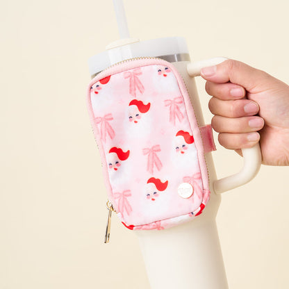 A hand holds a large cream tumbler with a handle, featuring The Darling Effect's Dear Santa Tumbler Fanny Pack—a pink zippered pouch with Santa faces and bows—attached. The background is plain light beige.