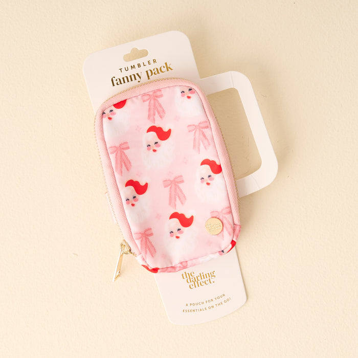 The Darling Effect's Dear Santa Tumbler Fanny Pack is a small, stylish pink pouch with Santa faces and bows, gold zipper, and attached to white packaging. Displayed on a light cream background.