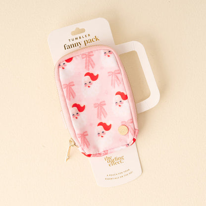 The Darling Effect's Dear Santa Tumbler Fanny Pack is a small, stylish pink pouch with Santa faces and bows, gold zipper, and attached to white packaging. Displayed on a light cream background.