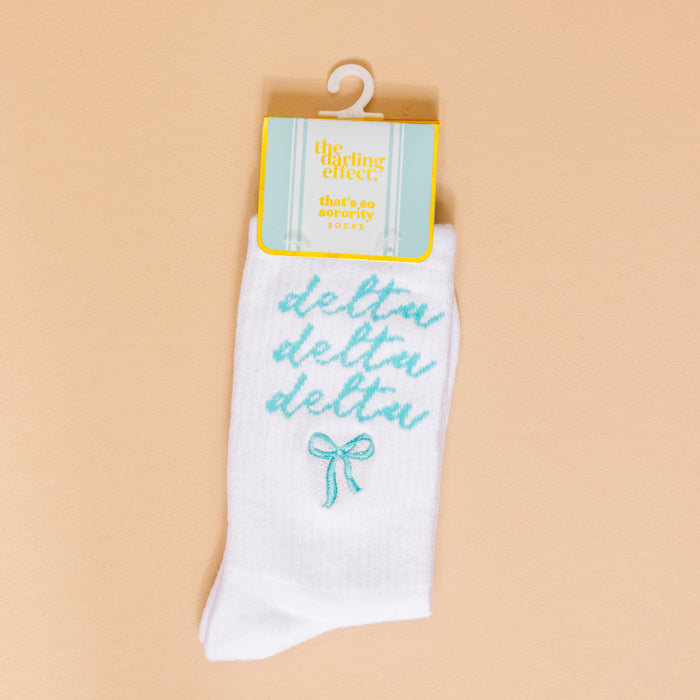 The Darling Effect’s Delta Delta Delta Embroidered Crew Socks feature teal cursive lettering and a teal bow—perfect for sorority gifts. Displayed on a peach background with retail tag.