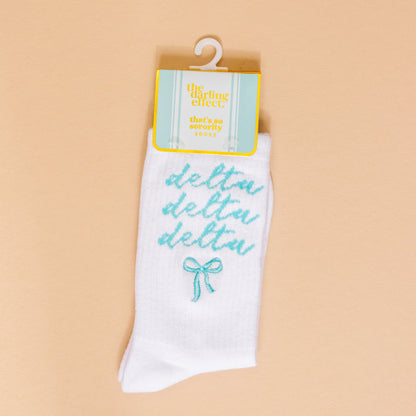 The Darling Effect’s Delta Delta Delta Embroidered Crew Socks feature teal cursive lettering and a teal bow—perfect for sorority gifts. Displayed on a peach background with retail tag.