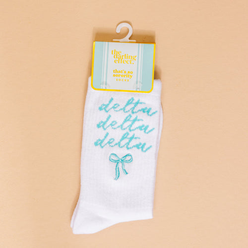The Darling Effect’s Delta Delta Delta Embroidered Crew Socks feature teal cursive lettering and a teal bow—perfect for sorority gifts. Displayed on a peach background with retail tag.