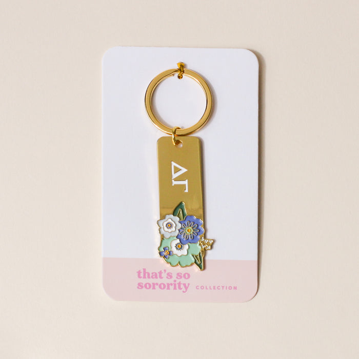 The Darling Effect’s Delta Gamma Enamel Keychain features gold-tone Greek letters with blue and white flowers, on packaging reading "thats so sorority COLLECTION"—a perfect gift for any Delta Gamma sister.