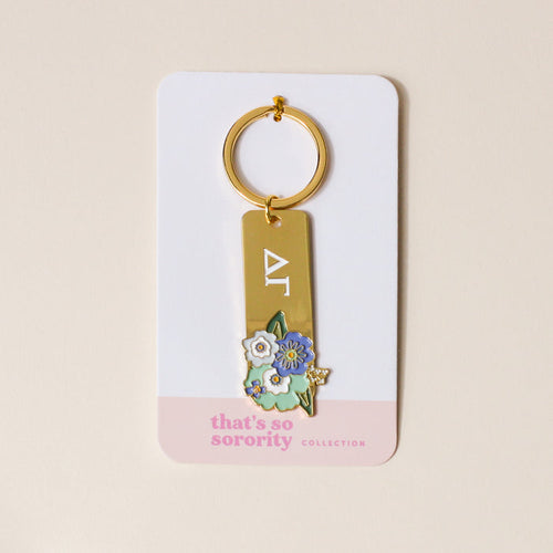 The Darling Effect’s Delta Gamma Enamel Keychain features gold-tone Greek letters with blue and white flowers, on packaging reading "thats so sorority COLLECTION"—a perfect gift for any Delta Gamma sister.