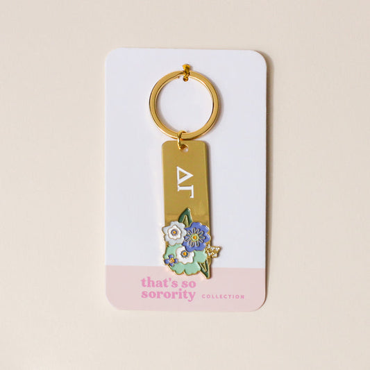 The Darling Effect’s Delta Gamma Enamel Keychain features gold-tone Greek letters with blue and white flowers, on packaging reading "thats so sorority COLLECTION"—a perfect gift for any Delta Gamma sister.