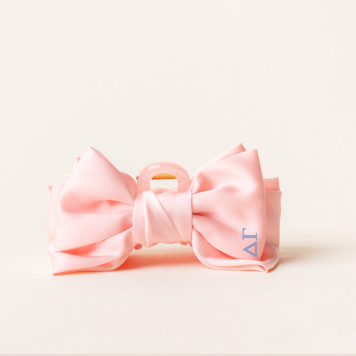 The Darling Effect Delta Gamma Satin Bow Claw Clip features light pink satin with blue Greek letters on the right tail—perfect for updos and styled against a cream background.