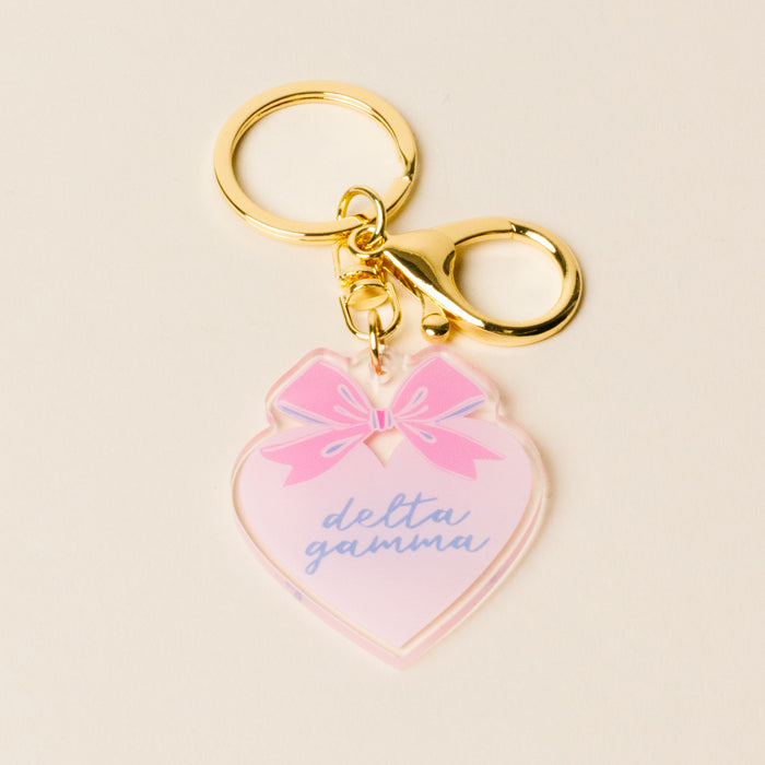 The Darling Effect Delta Gamma Sweetheart Bow Acrylic Keychain features a gold heart, pink bow, and blue "delta gamma" script—perfect as a sisterhood gift or cute bag accessory.