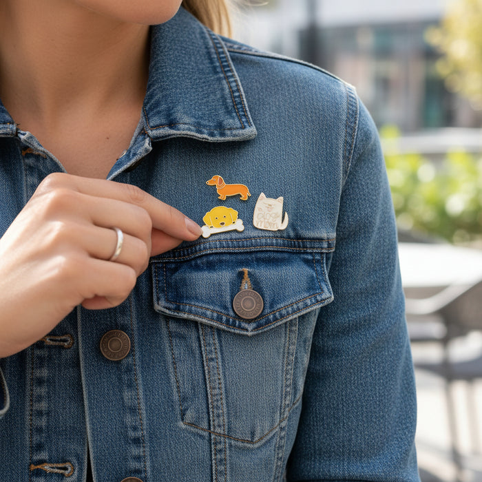 A person wearing a blue denim jacket displays The Darling Effect's Dog Days The Pin Pack—three collectible pins: a dachshund dog, a yellow smiley face, and a white cat. They hold one pin and wear a silver ring.