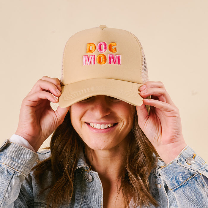 A smiling woman in a light denim jacket holds the brim of the-darling-effect's Dog Mom Trucker Hat, featuring colorful "DOG MOM" embroidery on beige, against a plain background.