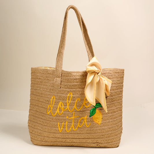 The Darling Effect Dolce Vita Straw & Sun Tote features "dolce vita" embroidered in yellow, a lemon design, and a pale yellow bow on one handle, displayed against a plain beige background.