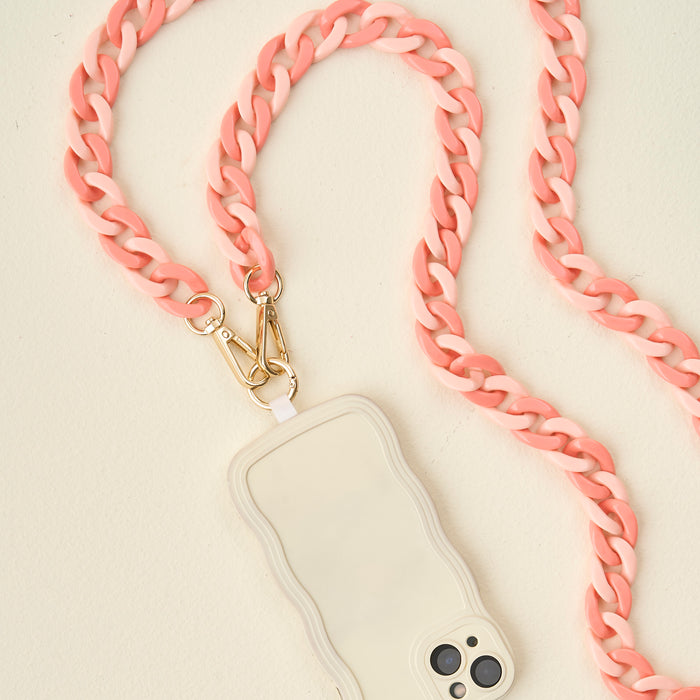 The Darling Effect's Duo Rose Hold the Phone Crossbody Chain features pink and white links with gold clasps, attached to a white smartphone in a wavy-edged case—an on-the-go accessory, displayed on a light beige surface.
