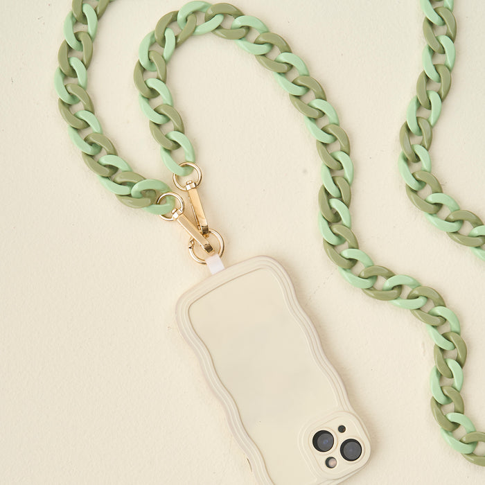 The Darling Effect's Duo Sage Hold the Phone Crossbody Chain, featuring chunky mint green and olive links with a gold clasp, is attached to a beige wavy-edged phone case resting on a light cream surface.