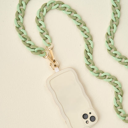 The Darling Effect's Duo Sage Hold the Phone Crossbody Chain, featuring chunky mint green and olive links with a gold clasp, is attached to a beige wavy-edged phone case resting on a light cream surface.
