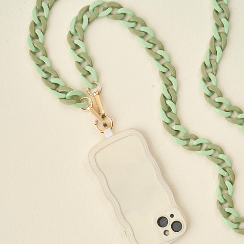 The Darling Effect's Duo Sage Hold the Phone Crossbody Chain, featuring chunky mint green and olive links with a gold clasp, is attached to a beige wavy-edged phone case resting on a light cream surface.