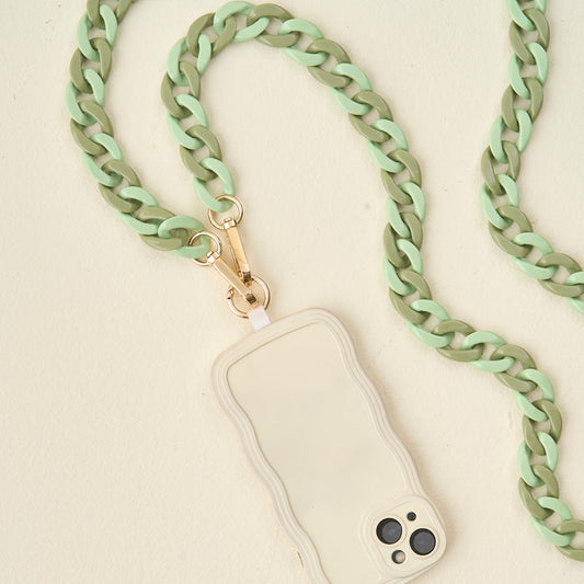 The Darling Effect's Duo Sage Hold the Phone Crossbody Chain, featuring chunky mint green and olive links with a gold clasp, is attached to a beige wavy-edged phone case resting on a light cream surface.
