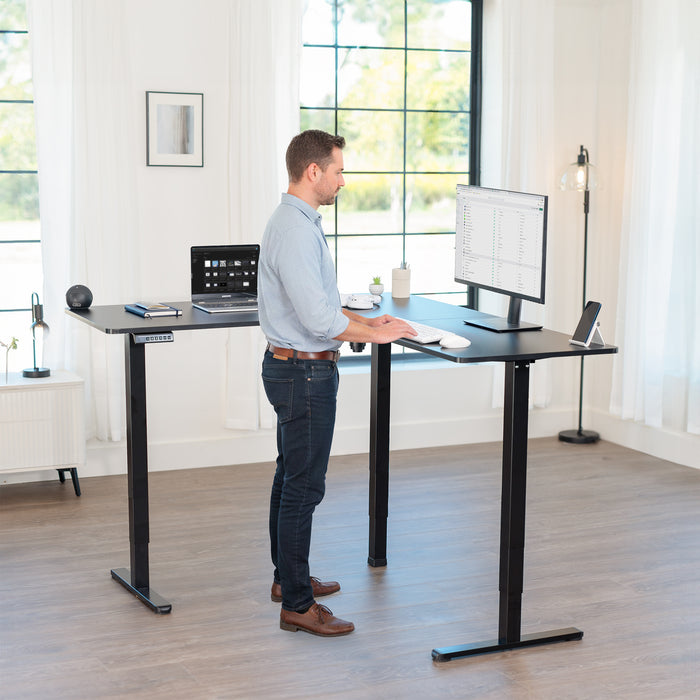 Man using a standing l-shaped desk in a bright room with large windows.