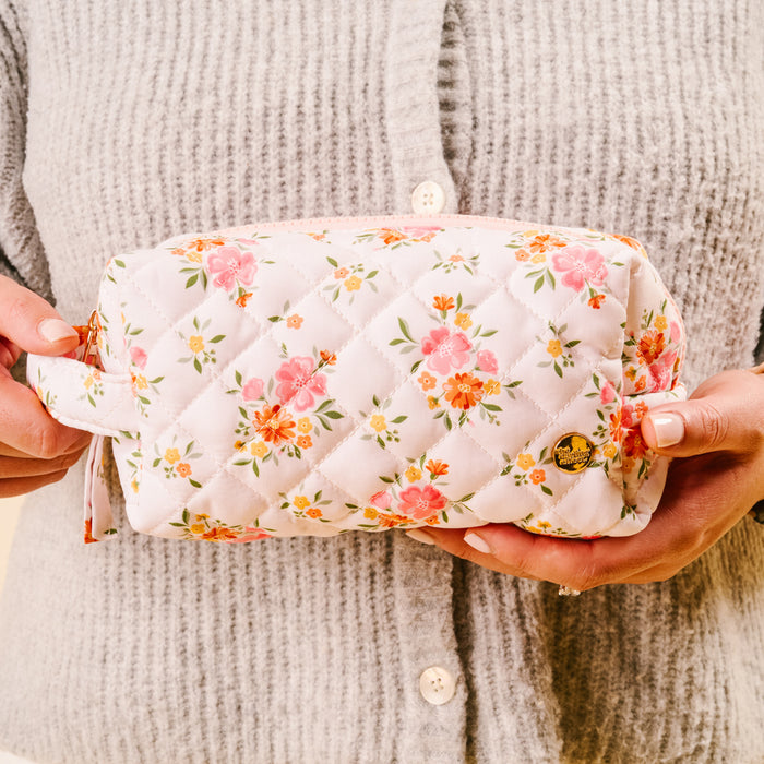 A person in a gray, ribbed cardigan holds the-darling-effect’s Endless Daydream Cream Essentials Quilted Cosmetic Pouch, featuring a pink and orange floral design, wipeable interior, and gold emblem on one corner.