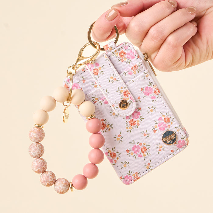 A hand holds The Darling Effect’s Endless Daydream Cream Keychain Wallet, a slim floral wallet with a zippered coin holder and a pink and beige beaded bracelet, showcased against a light background.