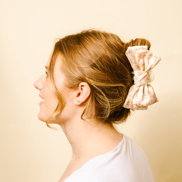 A woman with light brown hair in a bun, secured with the-darling-effect's Endless Daydream Cream Satin Bow Claw Clip. She smiles in a white top, viewed from the side against a light beige background.