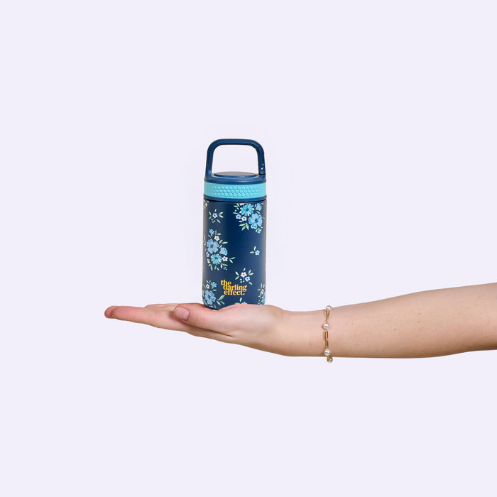 A hand with a bracelet holds The Darling Effect’s Endless Daydream Navy 6 oz Lil' Bitty Bottle, featuring a blue floral pattern and handle—perfect for hydration on the go against a plain light background.