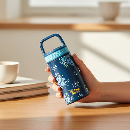 A hand holds The Darling Effect’s Endless Daydream Navy 6 oz Lil' Bitty Bottle, featuring a blue floral pattern and light blue lid. A coffee cup and books sit on a wooden table in the background. Perfect for hydration on the go.