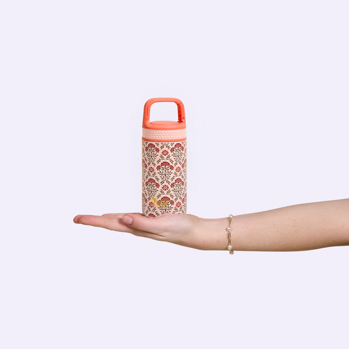 A hand with a bracelet holds the Everbloom Dusty Rose 6 oz Lil' Bitty Bottle by The Darling Effect, its pink pattern popping against a plain light background—the ideal compact travel companion for hydration anywhere.