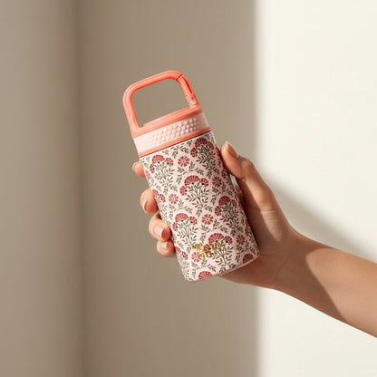 A hand holds The Darling Effect's Everbloom Dusty Rose 6 oz Lil' Bitty Bottle, featuring a floral design with a pink lid and handle against a softly lit neutral background—ideal for hydration on the go.