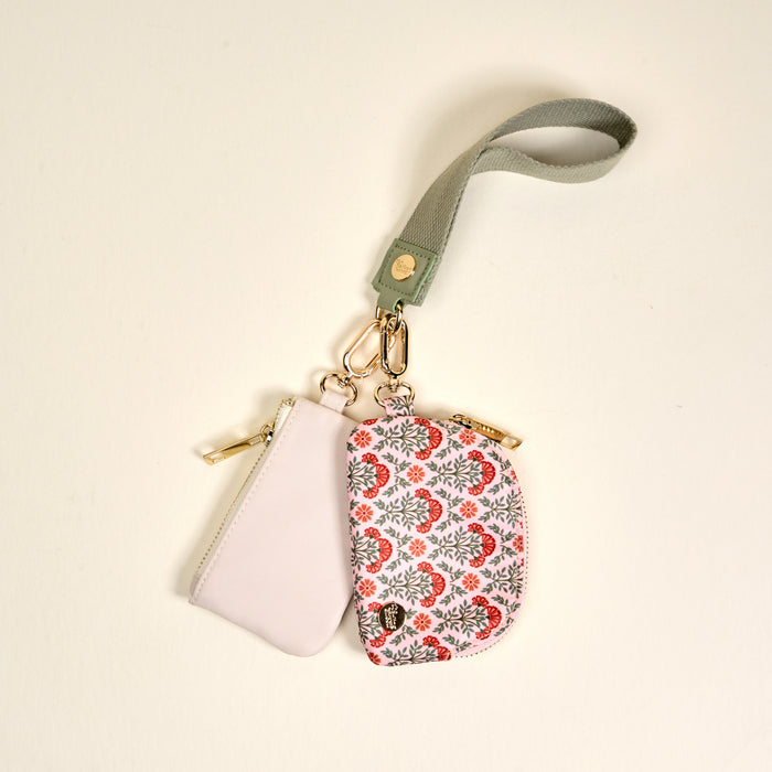 The Darling Effect’s Everbloom Dusty Rose Dynamic Duo Pouch Wristlet features two zippered pouches—one light pink, one white with red and grey florals—hanging from a green wristlet strap and secured with gold clasps for hands-free style.