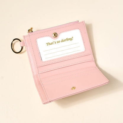 The Darling Effect’s Everbloom Dusty Rose Luxe Pocket Wallet, crafted from vegan leather, features a gold key ring, card slots, and an ID window with “That’s so darling!” in gold text—all shown on a light neutral background.