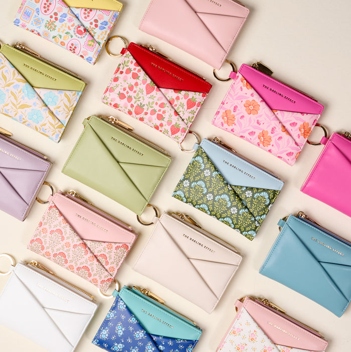 A flat lay features The Darling Effect's Everbloom Dusty Rose Luxe Pocket Wallet among colorful, compact wallets with criss-cross designs, floral patterns, or pastel shades—each with a gold zipper and key ring—arranged on a light background.