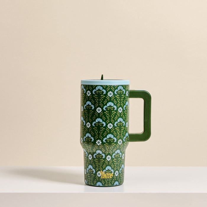 The Darling Effect Everbloom Forest Green 20 oz Lifestyle Flip Straw Tumbler with a large handle, blue lid, and insulated stainless steel body featuring a green and blue floral pattern rests on a white surface against a beige background.