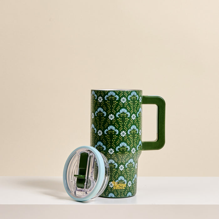 The Darling Effect Everbloom Forest Green 20 oz Lifestyle Flip Straw Tumbler features a white and blue floral pattern, large handle, insulated stainless steel body, and clear plastic lid with flip straw, shown on a white surface against a beige background.