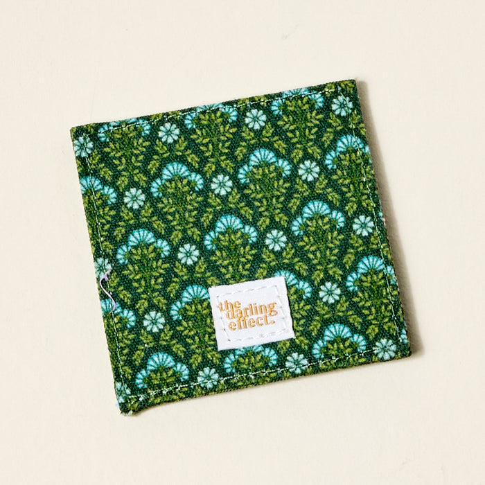 The Everbloom Forest Green Corner Keeper Fabric Bookmark by The Darling Effect is a square green fabric bookmark with a white and blue floral pattern and an orange-printed label—perfect as a unique book lover gift.