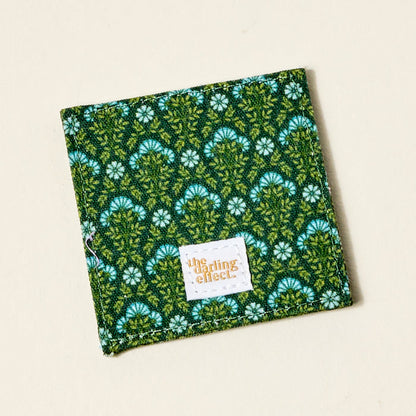 The Everbloom Forest Green Corner Keeper Fabric Bookmark by The Darling Effect is a square green fabric bookmark with a white and blue floral pattern and an orange-printed label—perfect as a unique book lover gift.