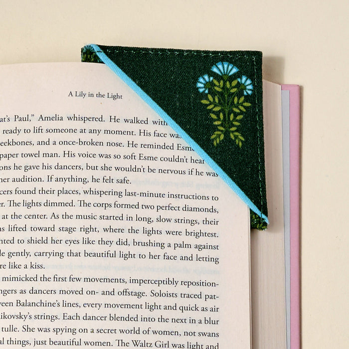 The Darling Effect's Everbloom Forest Green Corner Keeper fabric bookmark, embroidered with blue and yellow flowers, sits on the top right corner of an open book—an ideal gift for book lovers against a cream background.