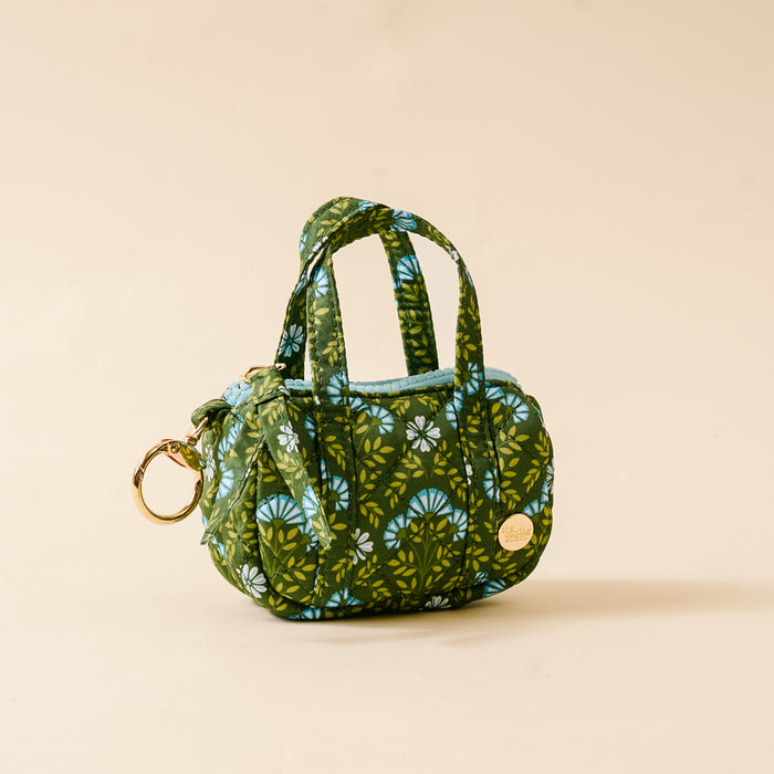 The Darling Effect’s Everbloom Forest Green Itty Bitty Duffle Bag Charm features quilted green fabric, blue and white florals, two short handles, a gold keyring, and a round gold tag, shown on a plain beige background.