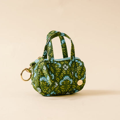 The Darling Effect’s Everbloom Forest Green Itty Bitty Duffle Bag Charm features quilted green fabric, blue and white florals, two short handles, a gold keyring, and a round gold tag, shown on a plain beige background.