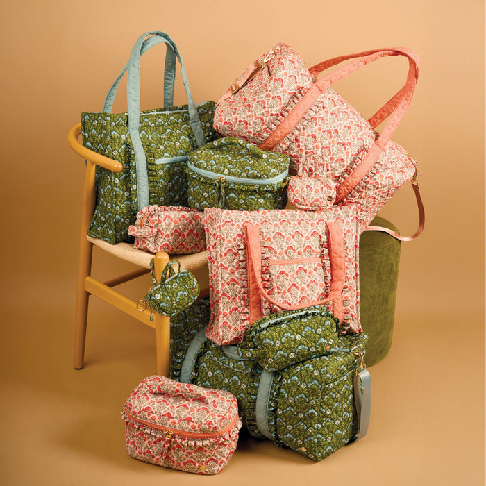 A variety of pink and green bags and accessories, including The Darling Effect’s Everbloom Forest Green Itty Bitty Duffle Bag Charm, are displayed on and around a wooden chair set against a beige background.