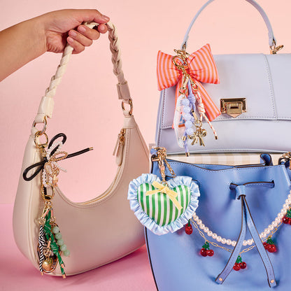 Three colorful handbags, each adorned with playful charms like bows, beads, hearts, cherries, and The Darling Effect's Everbloom Forest Green Itty Bitty Duffle Bag Charm, are displayed on a pink background. A hand holds a cream bag on the left.