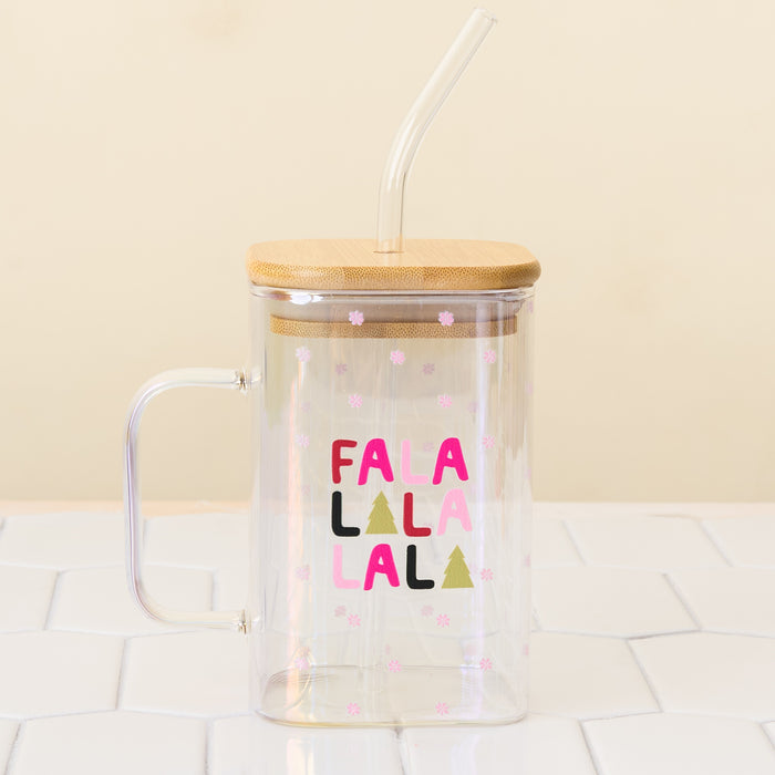 The Darling Effect Fa La La Square Glass Cup is a clear iridescent glass mug with a handle, bamboo lid, and glass straw. It features “FALALALALA” in colorful letters, Christmas trees, pink snowflakes, and is shown on a white tiled surface.