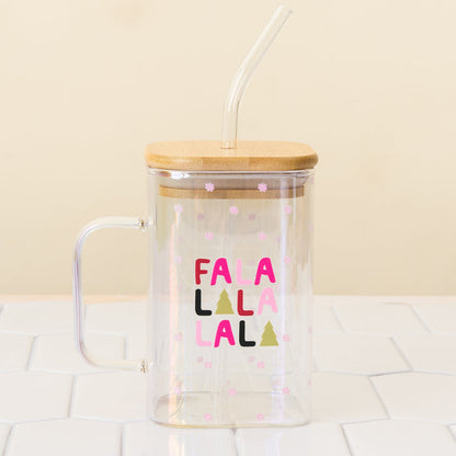The Darling Effect Fa La La Square Glass Cup is a clear iridescent glass mug with a handle, bamboo lid, and glass straw. It features “FALALALALA” in colorful letters, Christmas trees, pink snowflakes, and is shown on a white tiled surface.