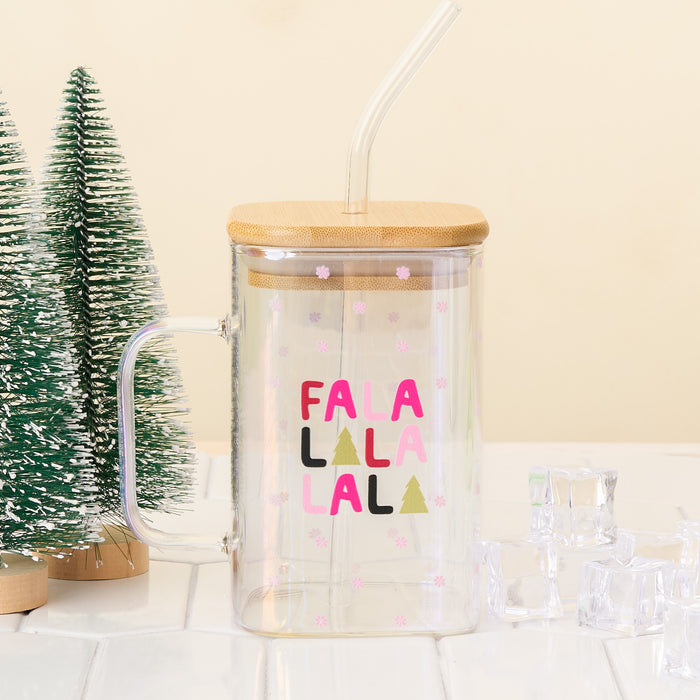 The Darling Effect’s Fa La La Square Glass Cup—a clear iridescent mug with bamboo lid, handle, straw, and pink snowflakes—sits on a white surface beside mini artificial pine trees and ice cubes.
