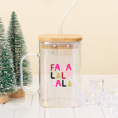 The Darling Effect’s Fa La La Square Glass Cup—a clear iridescent mug with bamboo lid, handle, straw, and pink snowflakes—sits on a white surface beside mini artificial pine trees and ice cubes.