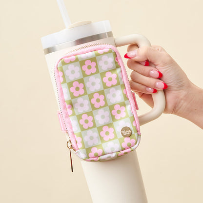 A hand with red French manicured nails holds a white tumbler with a straw, featuring The Darling Effect's Flower Check Green Tumbler Fanny Pack—a small, water-resistant pouch in a pink and green floral checkerboard pattern—on its handle.