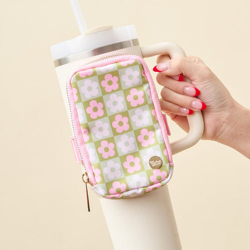 A hand with red French manicured nails holds a white tumbler with a straw, featuring The Darling Effect's Flower Check Green Tumbler Fanny Pack—a small, water-resistant pouch in a pink and green floral checkerboard pattern—on its handle.