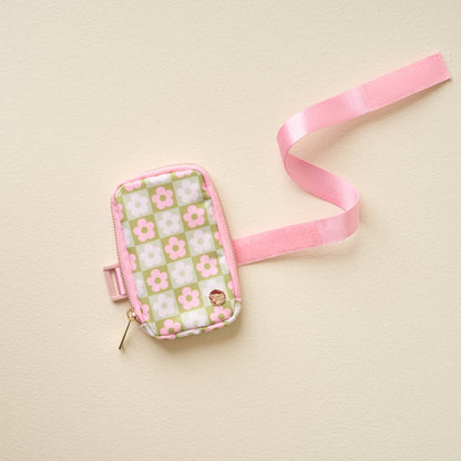 The Darling Effect’s Flower Check Green Tumbler Fanny Pack—a stylish pouch with pink strap and zipper, featuring white and pink flower checks on a green background—rests on a cream surface; perfect for water-resistant storage.